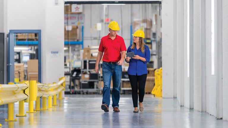 Two employees, a male and a female walking through a plant in deep conversation wearing yellow safety helmuts